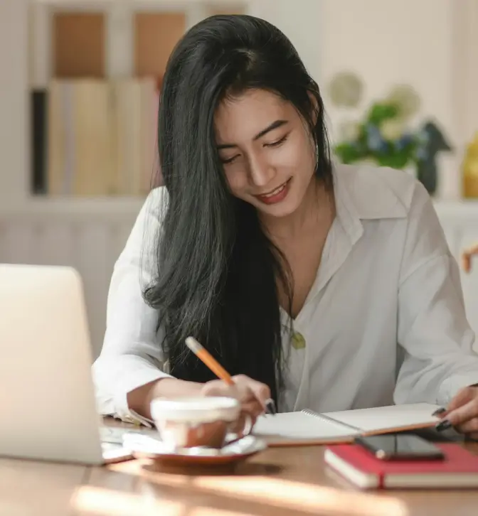 A portrait-style image of a young woman with long, dark hair working at a desk. She is wearing a white collared shirt and is looking down, smiling slightly as she writes with a pencil in a notebook. A laptop, a cup of coffee, and a red book are also visible on the wooden table, with a blurred, bookshelf-filled background suggesting a home office or study space.